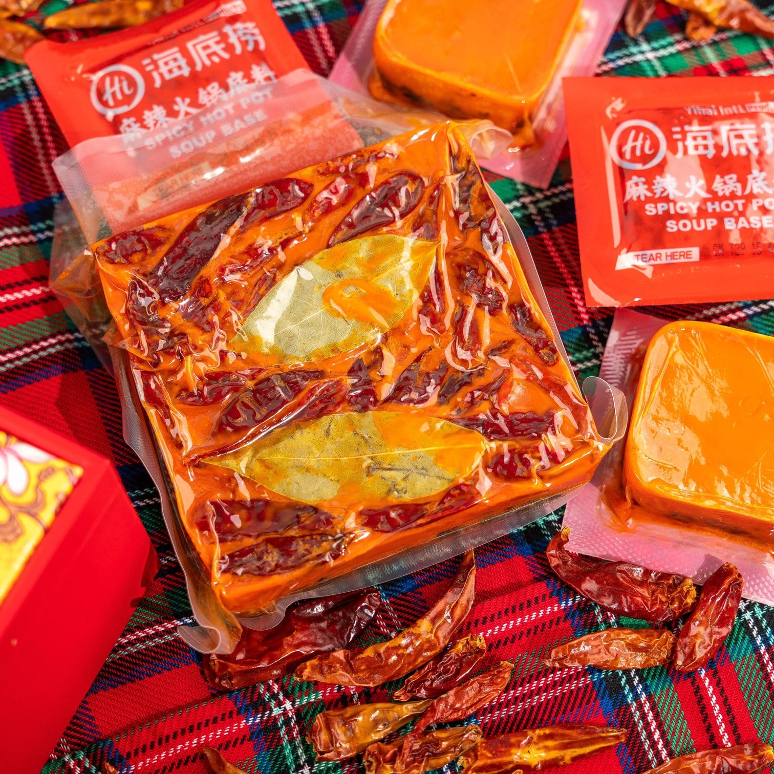 HAIDILAO Spicy Hot Pot Soup Base packages surrounded by dried chili peppers on a red plaid tablecloth