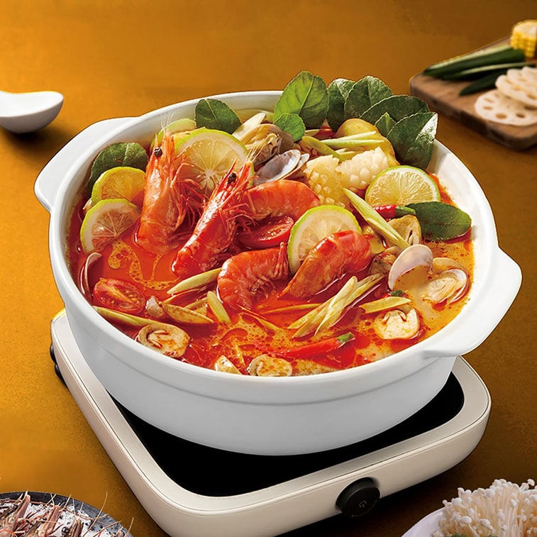 White ceramic hot pot on an induction cooker filled with tom yum soup, shrimp, clams, lime slices, mushrooms, lemongrass, and lettuce