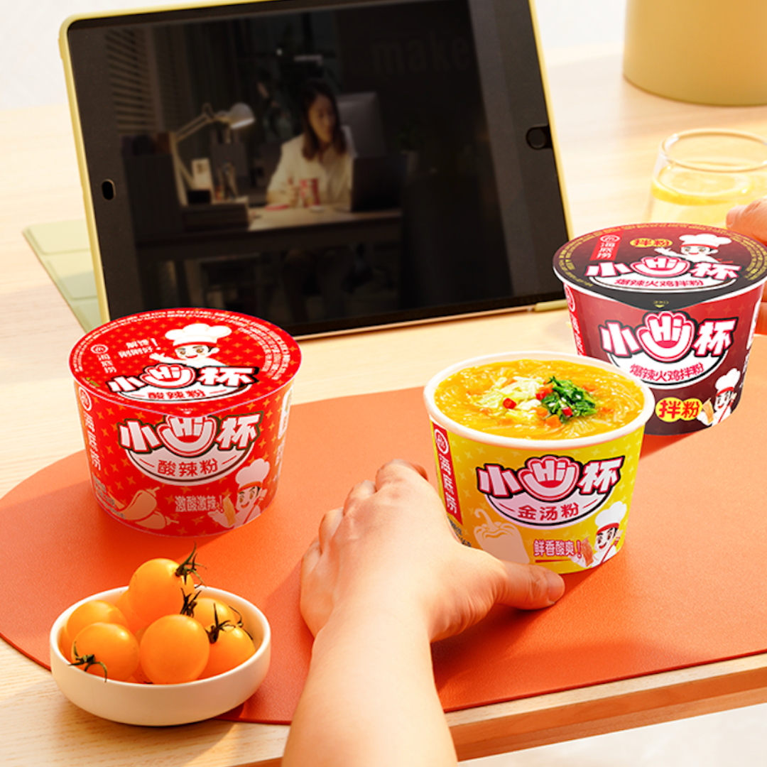HAIDILAO Mini Cup noodle varieties displayed on a picnic blanket with a tablet, hat, and fruit