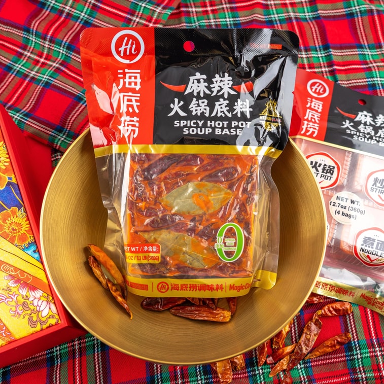 HAIDILAO Spicy Hot Pot Soup Base package displayed in a gold bowl on a red plaid tablecloth with dried chili peppers